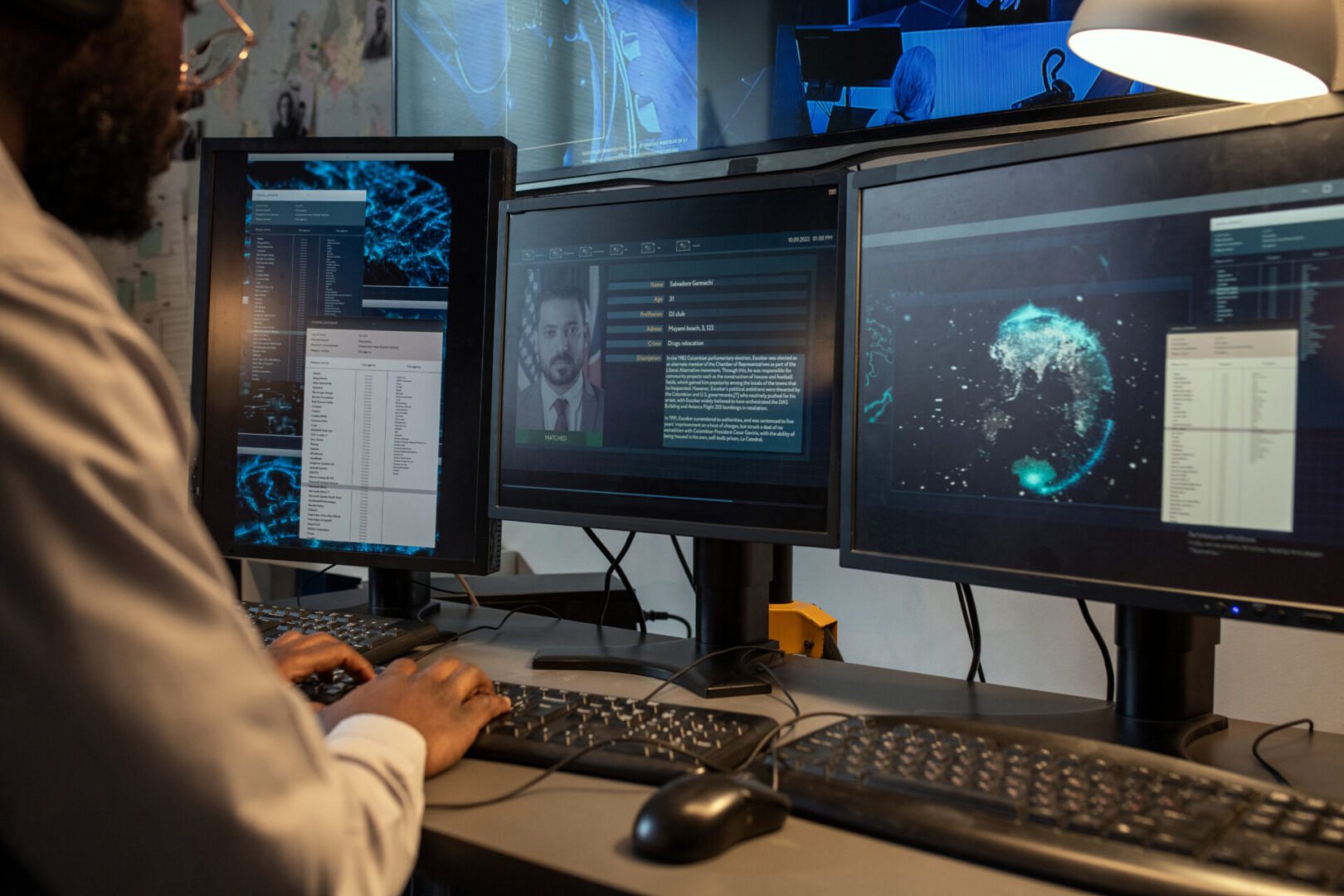 A person using a computer with multiple monitors displaying data, maps, and a portrait.